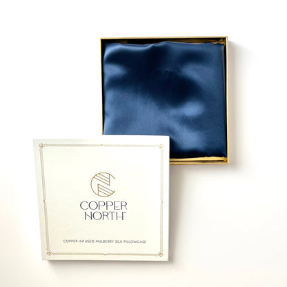 A Copper North brand pillowcase in deep  blue silk, shown with its gold-trimmed open box and the branded white lid in the foreground against a clean white background.