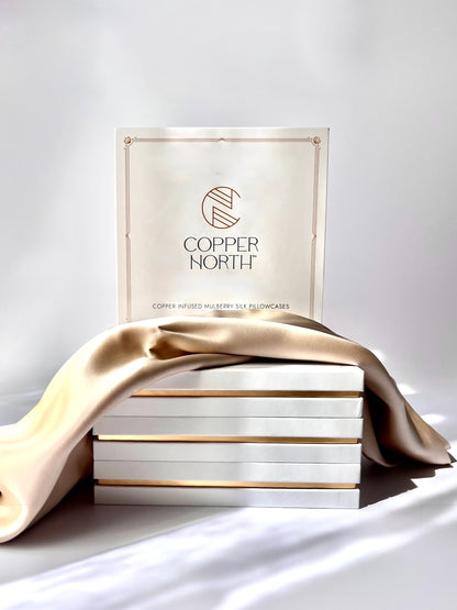 A stack of white boxes with gold trim and a cream-colored box labeled 'COPPER NORTH' stands behind draped champagne gold silk fabric on a white surface.