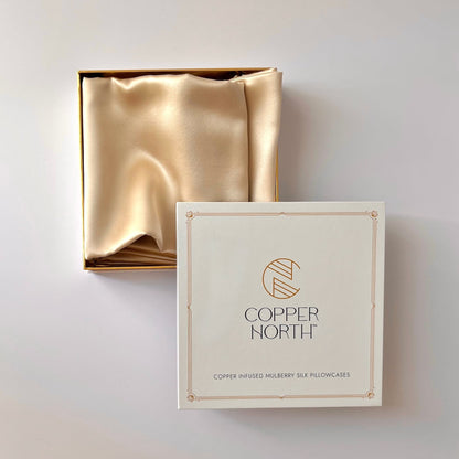 Two luxury pillowcases in a gold silk, shown folded inside their open matching box next to its cream-colored outer packaging. The packaging features the brand name 'COPPER NORTH' in elegant gold lettering.