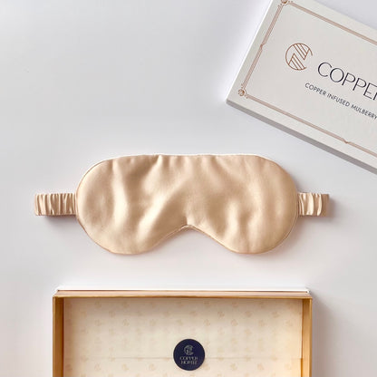 A champagne-gold silk sleep mask is displayed horizontally, with its packaging box partially visible at the top right and another open box at the bottom. 