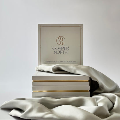 A stack of white boxes with gold-copper trim, featuring a square box standing upright in the back that reads 'COPPER NORTH' with a circular logo. A draped piece of light gray silk fabric covers the stack against a soft light gray background.