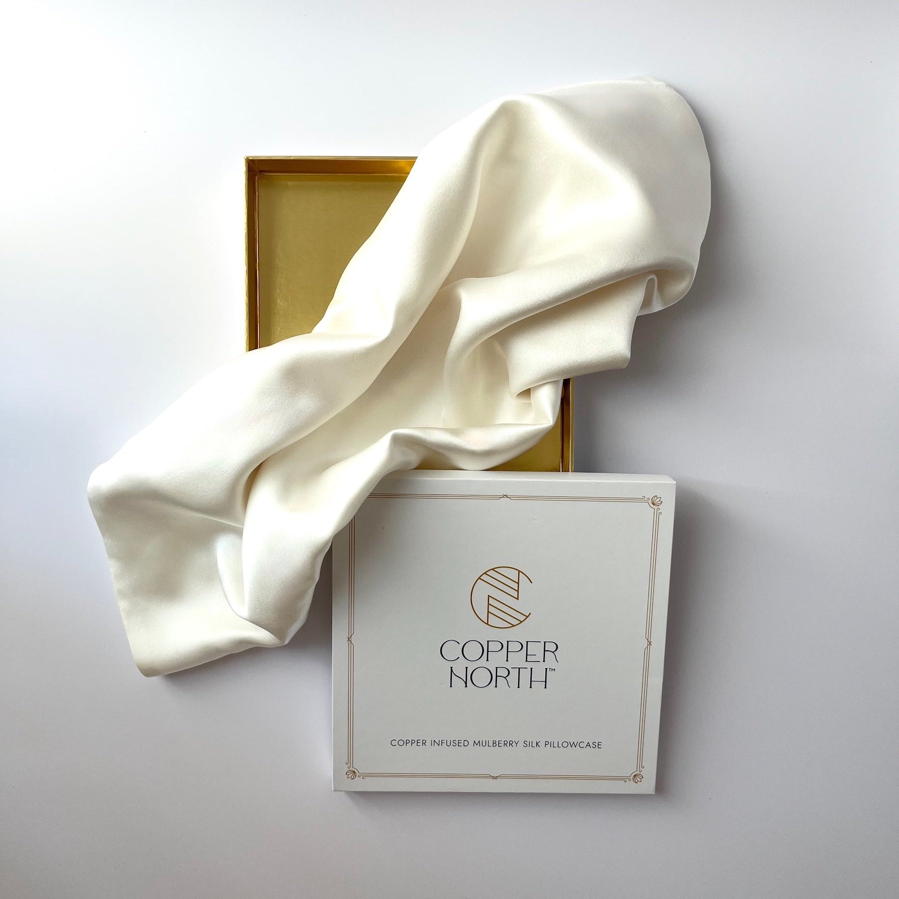 An white silk pillowcase draped over its white packaging box, which features the 'Copper North' brand logo, with a gold inner tray visible in the background against a light gray surface.