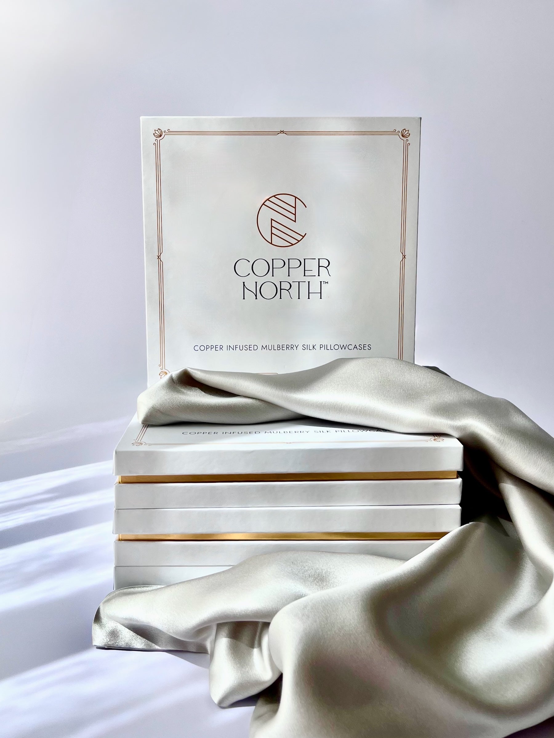 A stack of Copper North pillowcase boxes with a matching branded box standing behind them, draped with light gray silk on a clean white studio background.