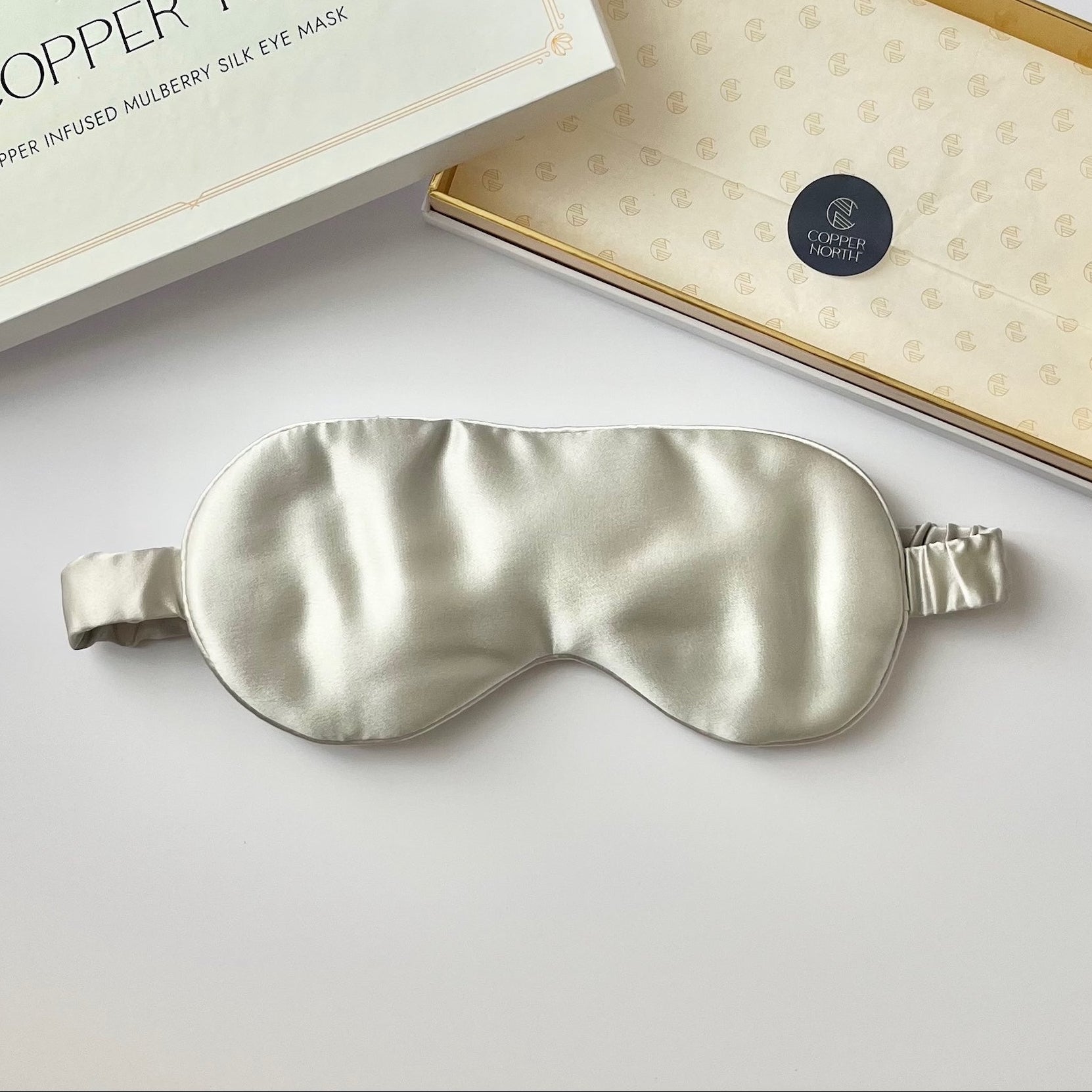 A silk sleep mask in a pale gray color, displayed next to its white packaging box on a clean white surface.