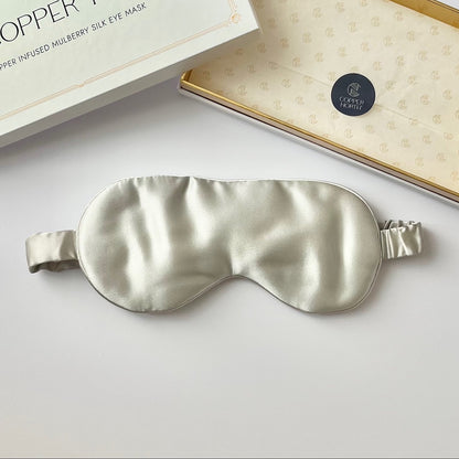 A silk sleep mask in a pale gray color, displayed next to its white packaging box on a clean white surface.