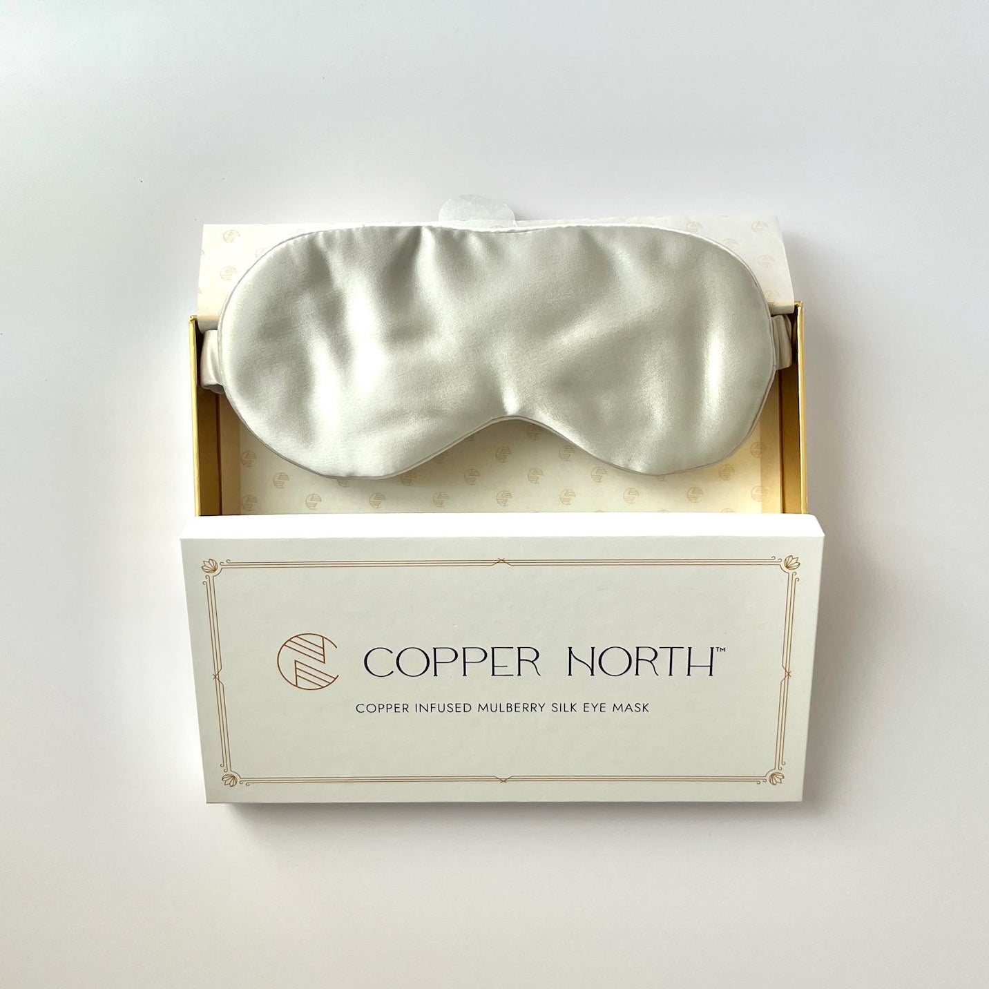 A pale gray silk sleep mask nestled in an open white gift box with gold trim, branded 'COPPER NORTH'. The box is set against a clean, light off-white background.