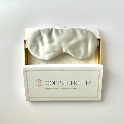 A pale gray silk sleep mask nestled in an open white gift box with gold trim, branded 'COPPER NORTH'. The box is set against a clean, light off-white background.