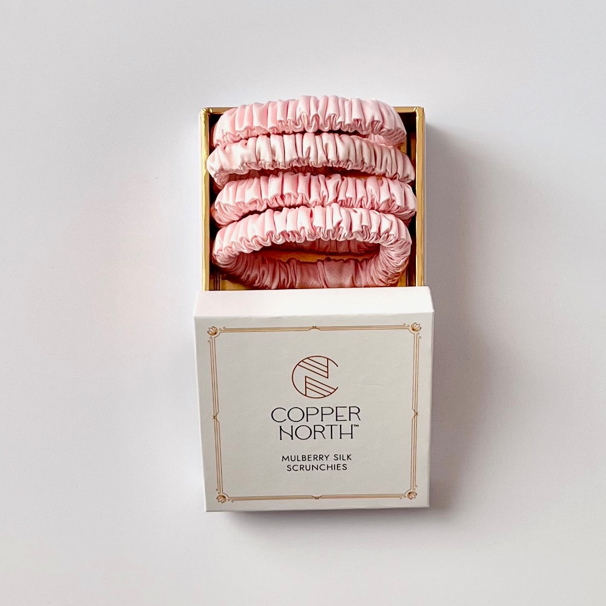 A set of four pink mulberry silk scrunchies displayed in an open gold-lined box above its white branded packaging box, set against a plain light gray background.