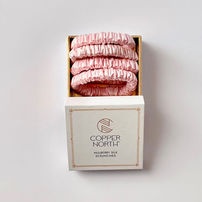 A set of four pink mulberry silk scrunchies displayed in an open gold-lined box above its white branded packaging box, set against a plain light gray background.