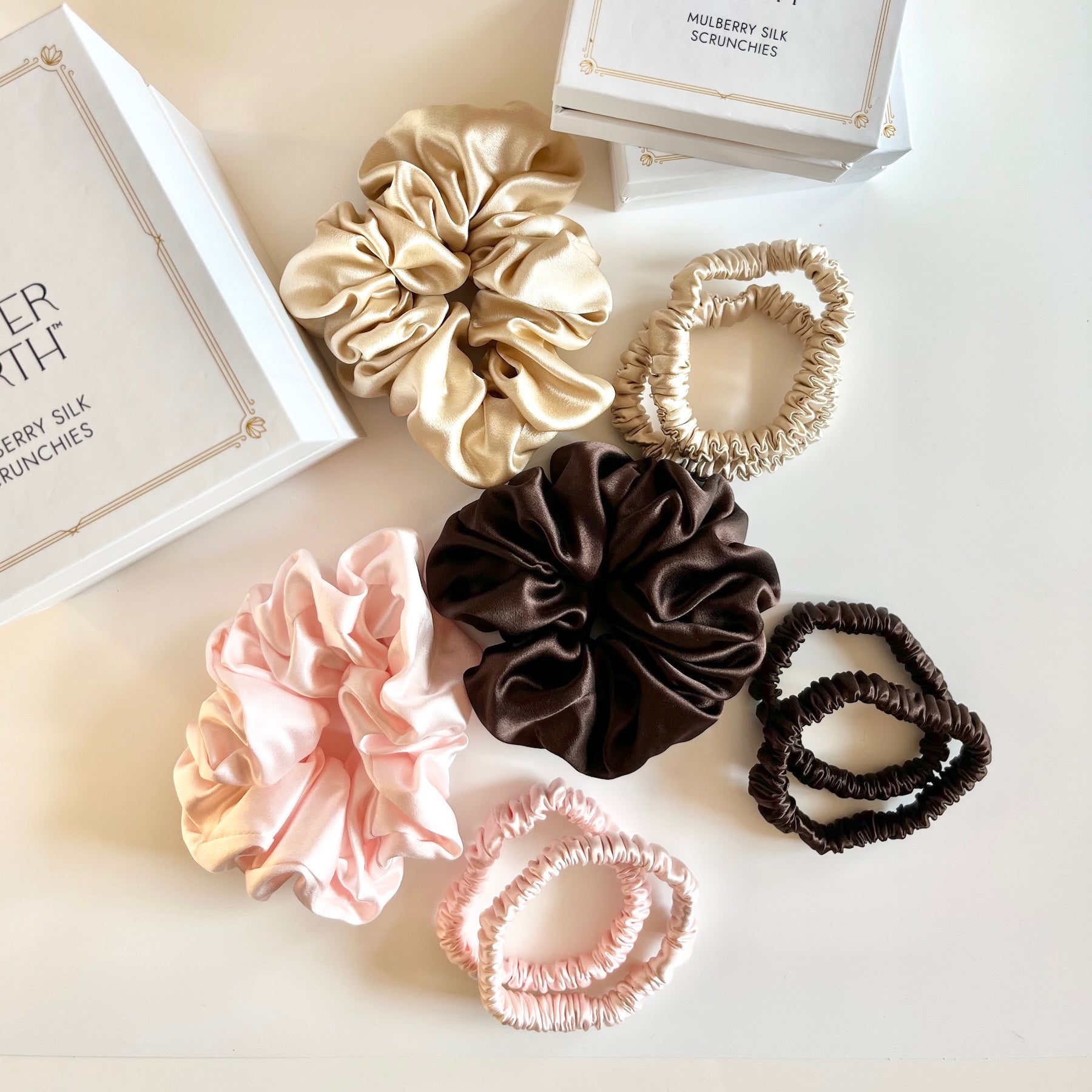 Nine silk scrunchies in assorted colors and textures are arranged on a white surface with Copper North gift boxes. The scrunchies include classic and sleek sizes in rose pink, gold, and chocolate brown.